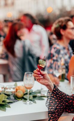 Een hand houdt een cocktailglas vast, met gasten op de achtergrond van een buitenfeest. Decor met bladeren op de tafel.